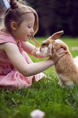 baby girl in a pink dress plays with a rabbit in a green meadow in summer. Funny friendship between a child and an animal during Easter.