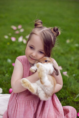 portrait baby girl in a pink dress plays with a rabbit in a green meadow in summer. Funny friendship between a child and an animal during Easter.