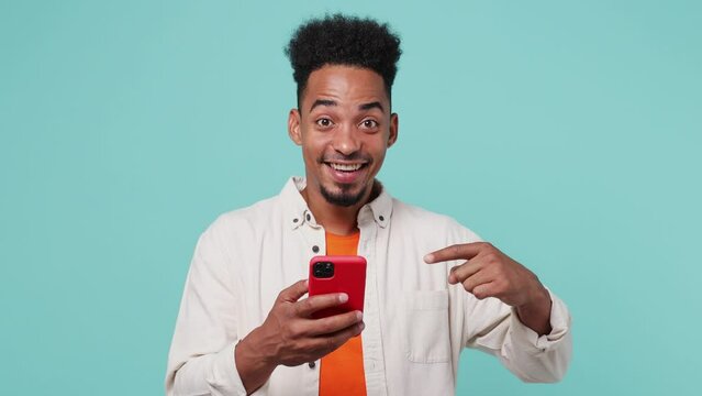 Young satisfied cheerful amazed man of African American ethnicity 20s he wear shirt t-shirt hold in hand use point index finger on mobile cell phone isolated on plain pastel light blue cyan background