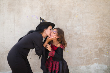 Fototapeta na wymiar Happy halloween. Young beautiful woman and girl dressed as witches on grey background. The woman is giving kisses to the girl on her face on the day of halloween party. Trick or treat.