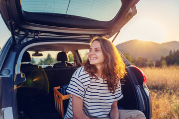 Young cheerful woman traveler sitting in open trunk of car enjoying sunset in nature