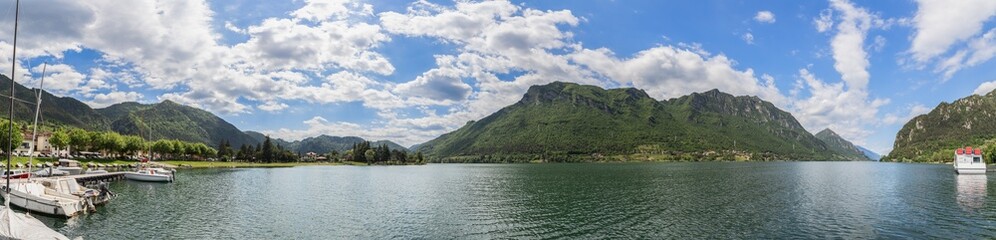 Maxi panoramic shot of glacier Lombard highland lake Idro (Lago d'Idro) surrounded by alpine wooded cliffs under piercing blue sky, several boats at a pier. Brescia, Lombardy, Italy