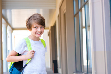 handsome happy little boy schoolboy with a backpack goes to school, stands at the school building	