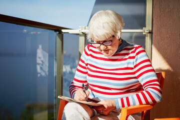 Elderly woman in glasses sitting on a balcony near the sea and looking at magazine, solving a...