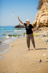 Senior woman in dark sunglasses feeds seagulls on the rocky beach. Happy life in retirement