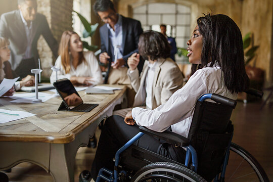 Office coworkers in the meeting room - main focus on African American woman with disabilities sitting on the wheelchair - business and disability lifestyle concept