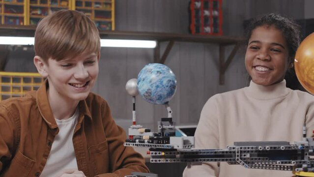 Cheerful Black girl and Caucasian boy working together on robotic solar system model doing high five