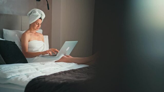 A young woman with a towel on her head working on a laptop in a hotel room during a business trip