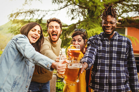 Cheers - multiracial group of happy people toasting joining beer mugs and laughing looking at the camera standing together outdoors in the countryside - people and alcohol lifestyle concept