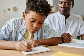 African little boy making notes in his notebook at table and doing homework with teacher