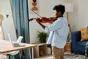 Rear view of African boy standing in his room and practicing to play violin during online lesson on...