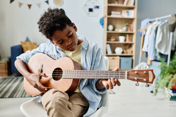 African boy sitting on chair and concentrating on his play in guitar, he remembering the notes during home lesson - obrazy, fototapety, plakaty