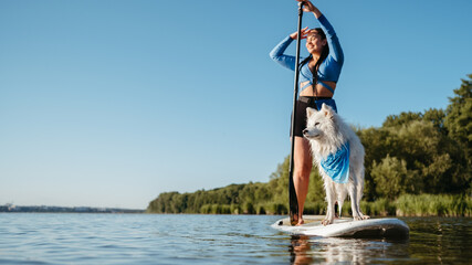 Happy Young Woman Enjoying Life on the Lake at Early Morning Standing on the Sup Board with Her Dog Snow-White Japanese Spitz