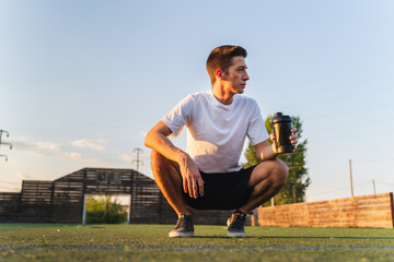 A young man is taking a break during the training to drink water and rest outside in nature during...