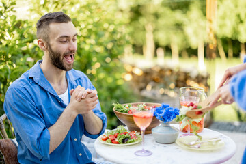 Excited man is waiting for lunch, watching enthusiastically as his wife serves healthy salad. Young family have a lunch at backyard during summer time