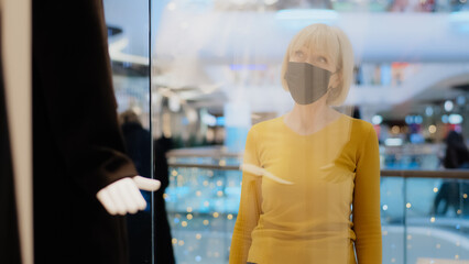 Mature woman in protective mask looking at showcase with mannequins in fashionable clothes middle-aged female shopaholic wants to update wardrobe admires dress sad because unobtainable expensive price