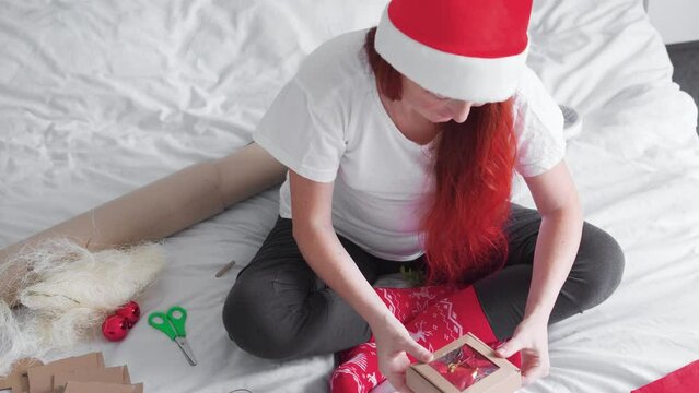 A girl receives and unpacks and opens a package from a kraft gift box with a red bow on a white background of Christmas and New Year decor. A red-haired woman with a red and white hat, a Christmas cap
