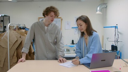 4K. A young female seamstress works with a client in a sewing workshop. They are looking at sketches of dresses