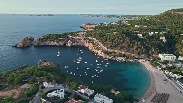 Sandy Cala Vadella Beach on Ibiza with rocky cliffs and houses on the hills. Many vessels and boats parked in a scenic small coastal Spanish village on the Balearic Islands