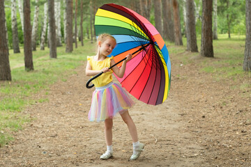 Happy funny girl holding rainbow umbrella. Cute Happy schoolgirl playing in rainy summer park. Kid walking in autumn shower. Outdoor fun by any weather