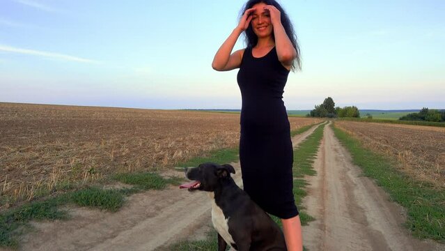 A beautiful young woman walks with a Staffordshire Terrier in a field. Happy free girl on a walk with a dog in nature. The concept of happiness, freedom, pets.