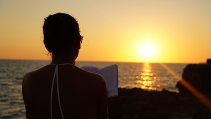 Fototapeta premium Silhouette of an elegant woman sitting on a rock in front of an idyllic sunset in sardegna italy, she breathes air of changes in her life and reads a book relaxed