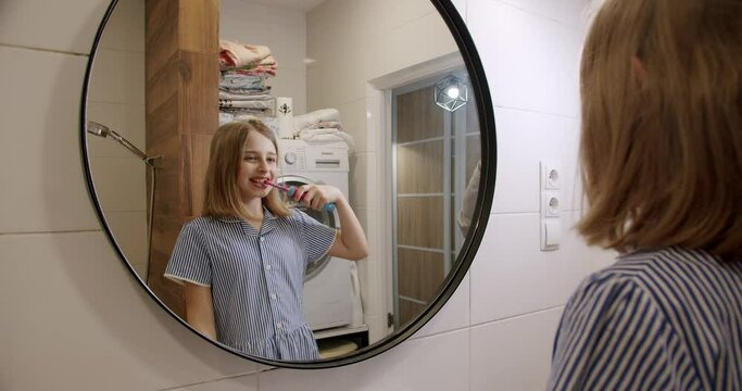 A teenage girl brushes her teeth with an electric toothbrush in the morning, in the bathroom and the modern house. Children daily healthcare routine Caucasian kid with white tooth looking at mirror.