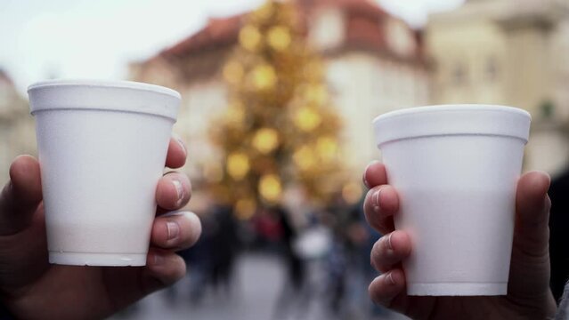 Hands and plastic cups with hot wine cheering, christmas tree, people and blurry lights in background, closeup view