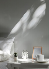 a white alarm clock stands on the bedside table in the morning sun next to the made-up bed