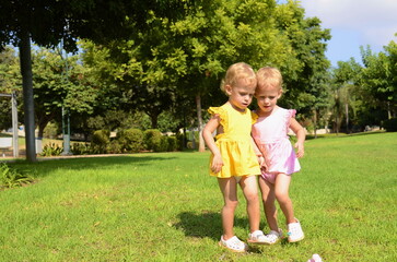 Fototapeta premium Cute twin girls, two years old. Blonde girls in yellow and pink dresses. The twins are walking in the park, the sisters are enjoying the summer.