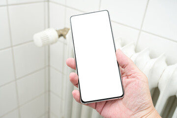 Mock up, hand holding a smartphone with blank white display in front of a heater radiator, concept...