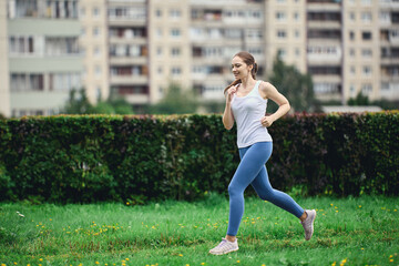 Fototapeta premium Woman in white t-shirt and leggings takes morning jog in residential area of city next to high-rise apartment building.