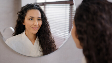 Fototapeta Close-up serious pensive young hispanic woman looking in mirror in bathroom contemplating future cute curly brunette girl in bathrobe admires reflection natural beauty daily morning routine concept obraz