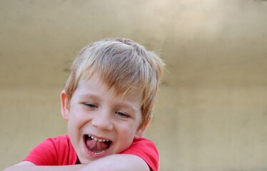 The blond boy has his eyes down and is smiling with his mouth wide open. A joyful five-year-old boy is fooling around. 