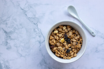 Bowl of homemade granola cereal with ceramic spoon on white marble table background. Top view, flat lay, copy space. Healthy eating concept. 