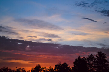 Morning glow illuminates the clouds on a beautiful colorful sky above the silhouettes of trees at dawn