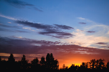 The morning glow illuminates the clouds on a beautiful colorful sky above the silhouettes of trees and a pillar at dawn