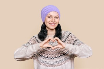 Young teenage female in hat sweater showing heart gesture with hands on brown background