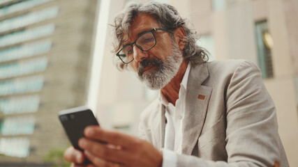 Fototapeta na wymiar Mature businessman with beard in eyeglasses wearing gray jacket holds cellphone in his hand and looks around pondering
