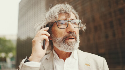 Clouse up, mature businessman with beard in eyeglasses wearing gray jacket is using cell phone. Middle aged manager talking on smartphone while sitting outside office