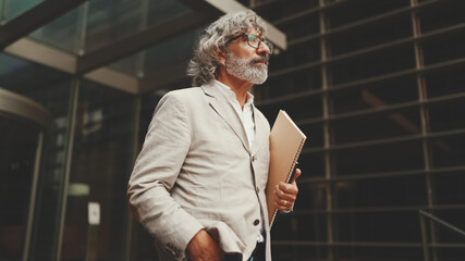 Mature businessman with beard in glasses wearing gray jacket holding folder with business documents is walking down the stairs leaving the business center