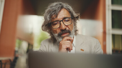 Fototapeta premium Clouse up, mature businessman with beard in eyeglasses wearing gray jacket sits on cafe. Middle aged manager successful man user on laptop pc computer sit at cafe outdoors