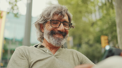 Positive middle-aged man with gray hair and beard wearing casual clothes sits on bench and uses mobile phone. Mature gentleman in eyeglasses writes message on smartphone