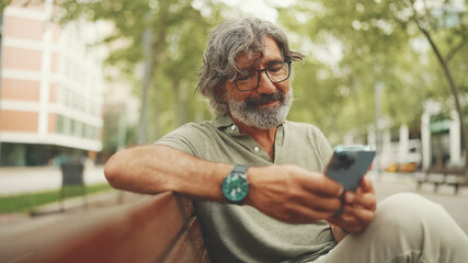 Positive middle-aged man with gray hair and beard wearing casual clothes sits on bench and uses mobile phone. Mature gentleman in eyeglasses writes message on smartphone