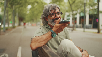 Clouse-up, smiling middle-aged man with gray hair and beard sits on bench and uses mobile phone....