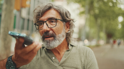 Clouse-up, smiling middle-aged man with gray hair and beard sits on bench and uses mobile phone....