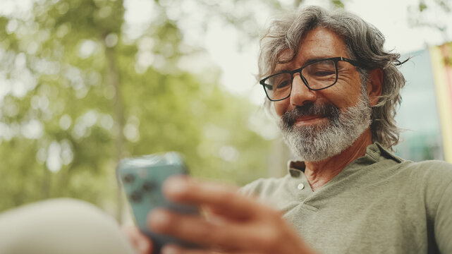 Positive middle-aged man with gray hair and beard wearing casual clothes sits on bench and uses mobile phone. Mature gentleman in eyeglasses writes message on smartphone