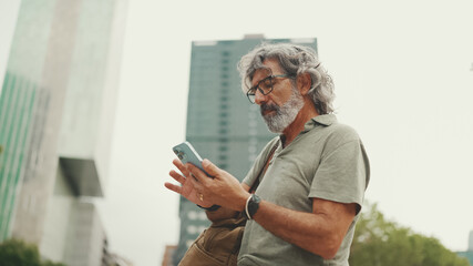 Friendly middle aged man with gray hair and beard looking at map trying to find his way using his mobile phone. Mature gentleman in eyeglasses using map app in cellphone outdoors