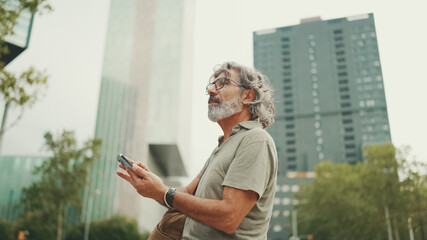 Friendly middle aged man with gray hair and beard looking at map trying to find his way using his mobile phone. Mature gentleman in eyeglasses using map app in cellphone outdoors