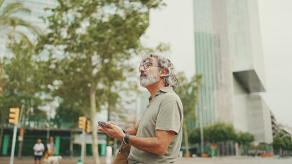 Friendly middle aged man with gray hair and beard looking at map trying to find his way using his mobile phone. Mature gentleman in eyeglasses using map app in cellphone outdoors